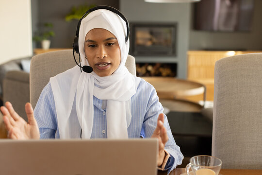 Adult woman wearing white hijab, sitting in living room, speaking on headset with laptop and cup