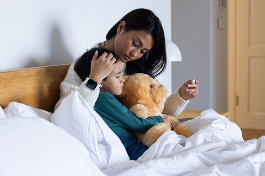 Asian mother and child sitting on bed, child clutching teddy bear while mother holding white device