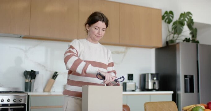 Adult woman in striped sweater, cutting tape with scissors on new box at counter, checking contents