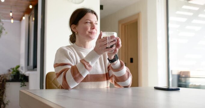 Adult woman in striped knit lifting mug to mouth at table while sunlight drawing gaze, phone