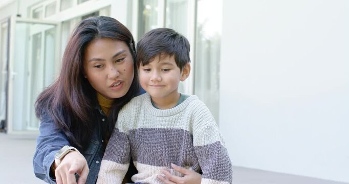 Asian woman pointing at schoolboy on bench by glass doors with smartwatch, guiding attention