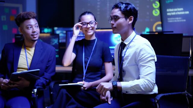 Black and Asian developers bump fists past a laughing Latina colleague, then chat cheerfully as the Asian man points upward in a dark AI lab with glowing screens