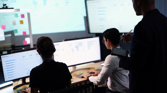 Latina woman in glasses turns to look up at a standing colleague with tablet while Asian programmer types and checks world map data on a large monitor in dark office