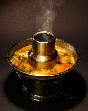 Steaming seafood soup with vegetables, herbs, and chili rests in a brass chimney hot pot over a tabletop burner, with a rich golden broth and rising steam against a dusky, moody restaurant background.
