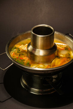 Steaming seafood soup with vegetables, herbs, and chili rests in a brass chimney hot pot over a tabletop burner, with a rich golden broth and rising steam against a dusky, moody restaurant background.
