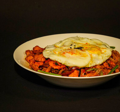 Thai drunken noodles stir-fried with meat, vegetables, and green onions, topped with a fried egg and chili flakes, served in a white bowl against a dark background, a savory restaurant dish for menus.