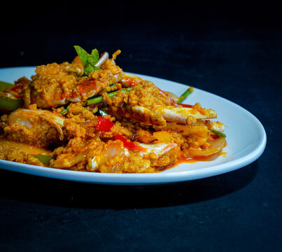 Crab pieces and claws coated in golden yellow curry sauce with vegetables, peppers, and herbs, served on a white plate against a dark background, showcasing a savory seafood dish for restaurant menus.