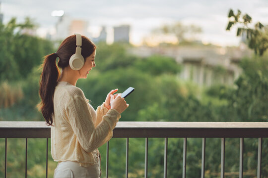 Smiling young Asian woman wearing wireless headphones and using smartphone outdoors by railing, enjoying digital connectivity and mobile entertainment with green nature and blurred city skyline in bac