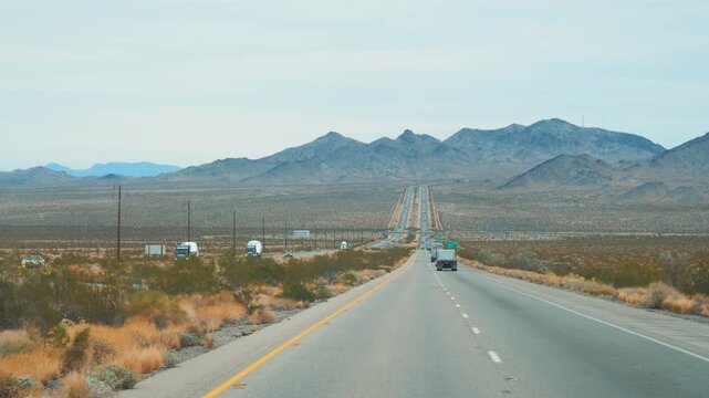 Vehicle and truck driving along straight asphalt desert highway toward rugged mountains, capturing vast arid landscape of american southwest during road trip. Pov driving on modern interstate speedway