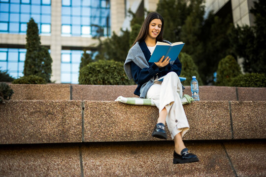 Modern Gen Z female college student sitting outside university with a book.