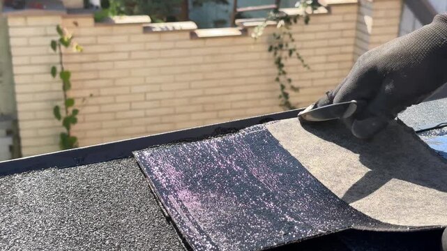 Roofer trims flexible bitumen shingles along the edge of a roof