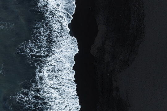 Aerial top down perspective shows white sea foam and breaking ocean waves meeting the volcanic black sand coastline near Vik in southern Iceland.