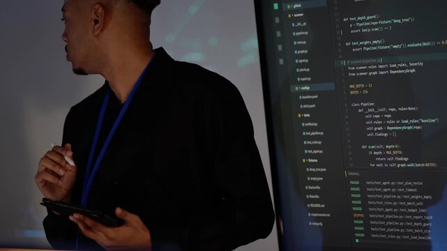 Close-up of a young Black AI engineer in a black shirt and lanyard pointing a stylus at Python code on a giant screen, then turning away and raising the pen to his chin in thought