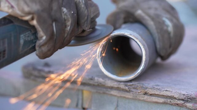 Gloved hands guide an angle grinder along the open rim of a metal pipe, sending sparks from the abrasive disc during edge preparation for metal fabrication and pipework