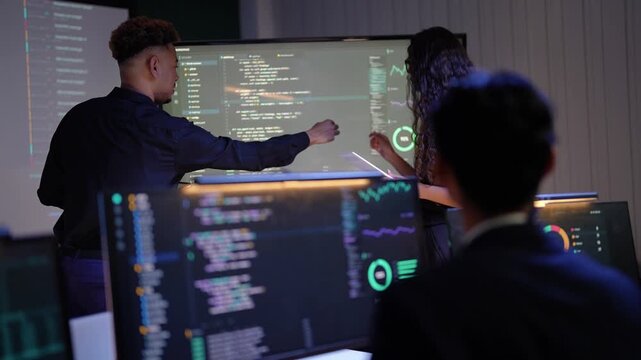Black engineer in a black shirt turns his back to the room and writes on the interactive code display beside a Latina coworker with a tablet, watched by an Asian man in the foreground