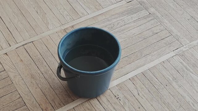Plastic bucket collecting leaking water drops on parquet floor during indoor house roof damage emergency problem