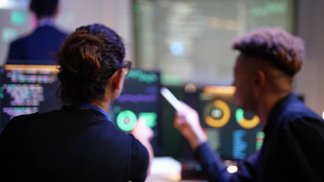 Latina coder points at a dashboard monitor and a Black teammate answers with his stylus as they review AI metrics side by side, an Asian presenter softly blurred at the wall screen