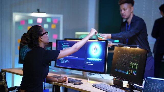 Latina developer at her desk touches a glowing AI logo screen with a stylus, then reaches out to tap it again while a Black teammate with a tablet stands talking behind her