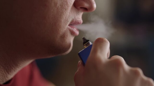 Closeup of person inhaling from blue electronic cigarette, bring vape pen to lips and exhale thick cloud of white vapor. Unhealthy habit and addiction concept, close-up, cropped shot, slow motion.
