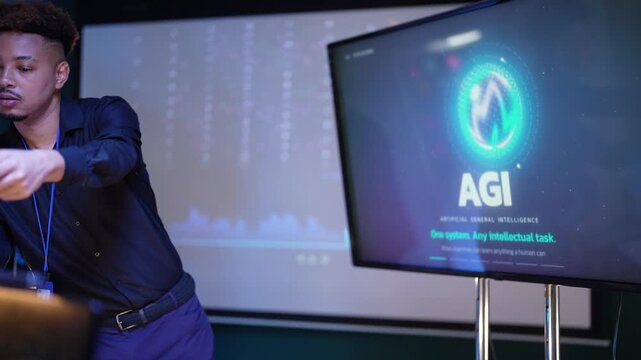 Black speaker reads his tablet, leans forward to hand a note to the audience, then talks with a raised stylus beside a glowing AI logo slide in a dim training room