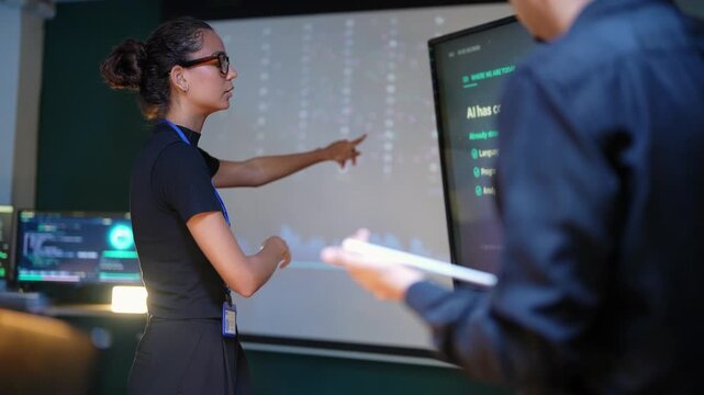 Latina developer raises a finger toward an AI capabilities slide, then reaches out to tap the large touchscreen while a Black teammate in the foreground holds his tablet and watches