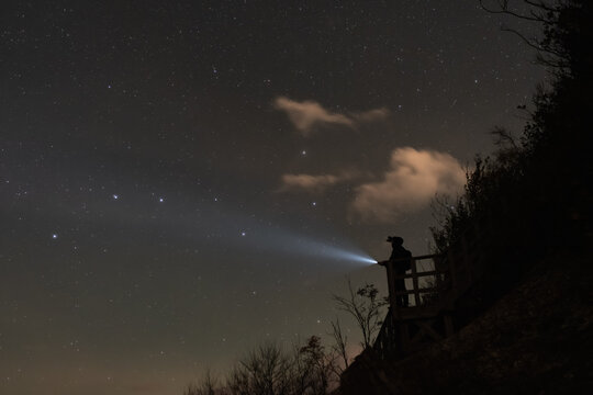 Man with flashlight standing on wooden viewpoint railing at night pointing light beam into starry sky.