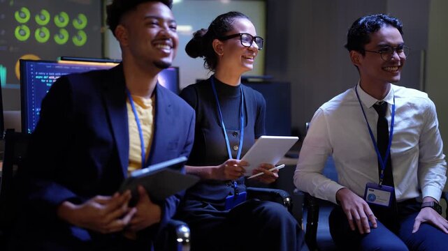 Three young tech colleagues seated side by side with tablets laugh together in a dark AI lab, a Black man in a navy blazer, a Latina woman in glasses and an Asian man in a white shirt