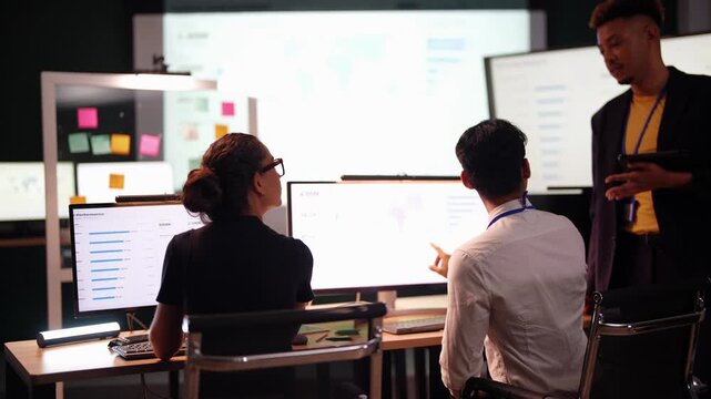 Asian developer in a white shirt turns from his bright monitor to talk to a Black colleague who stands taking notes on a tablet, the Latina coworker in a bun watches the screen