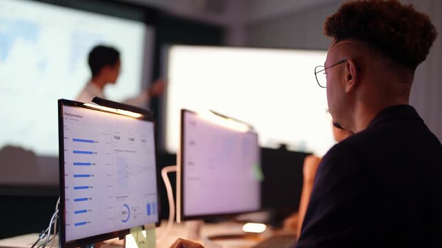 Over the shoulder of a Black developer at a performance analytics monitor while an Asian presenter gestures at the projected map and charts and a teammate raises a hand