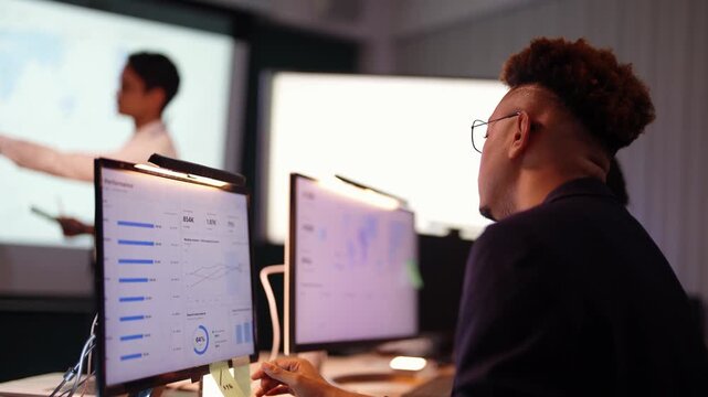 Black developer reaches out and points at bar charts on his monitor while an Asian presenter writes on a tablet at the projected map, a Latina teammate seated at right
