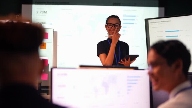 Smiling Latina woman with a hair bun taps notes on a tablet, laughs, then turns in profile toward the bright growth chart screen behind the blurred heads of two seated colleagues.