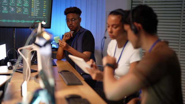 Black man adjusts his glasses and studies a green sticky note while a Latina woman with a tablet and a blurred Asian man in a headset lean toward the desk beneath the dashboard.