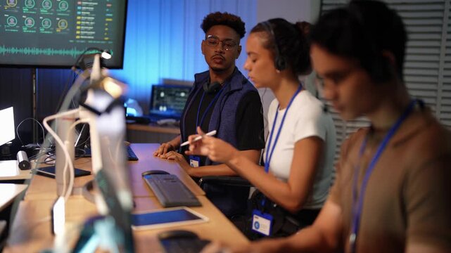 Latina woman with headset rests her chin on her hand, then takes notes with a stylus as a Black colleague holds a green sticky note, a blurred Asian man in a brown polo in front.