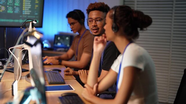 Latina woman in headset types at her screen, then the focus pulls past her to a Black developer and an Asian man in brown polo typing side by side at a long row of monitors.