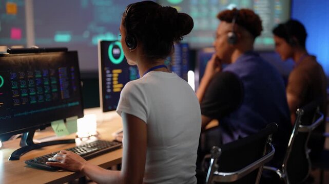 Over the shoulder of a Latina woman in a white tee who types, then rests her chin on folded hands studying the big screen, Black and Asian colleagues with headsets beside her.