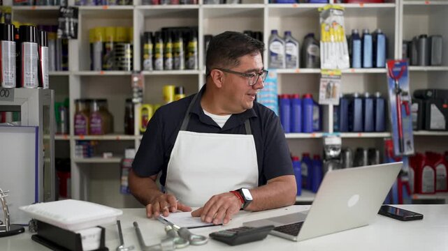 Hardware store manager writing notes while working behind counter with laptop and shelves