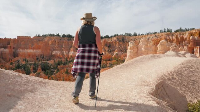 Active female hiker walking along scenic ridge with trekking poles while admiring famous red rock hoodoos at bryce canyon national park on sunny day
