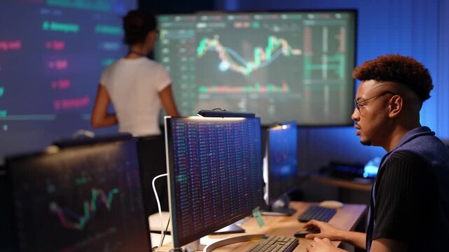 Latina analyst with a tablet reviews stock candlestick charts on a wall screen as a Black developer in a blue vest types and an Asian colleague reaches over to point at his monitor