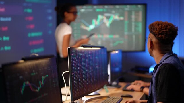 Black developer with glasses and a fade haircut focuses on his trading monitor then turns toward a Latina analyst checking a stock chart on her tablet beside the large wall screen