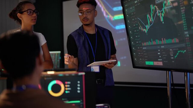 Black presenter in a blue vest points his stylus at a candlestick chart on the screen and then explains to the audience, a Latina colleague beside him, an Asian man watching from behind
