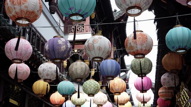 Rows of paper lanterns in pink, teal, purple and orange hang above a lane in old-town Shanghai near Yu Garden, traditional wooden eaves behind, bright sky, China