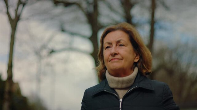 Thoughtful Caucasian older woman under bare trees and cloudy sky