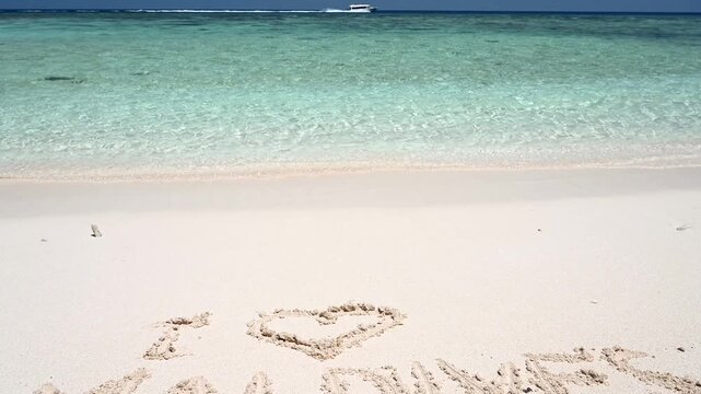 I love Maldives written in white sand on a tropical beach. Camera tilts up to reveal clear turquoise ocean water and a boat traveling on the horizon. Summer vacation and travel destination concept