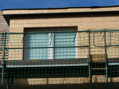 a building under construction, showing a newly installed window set into light wood cladding, protected by construction scaffolding with a green safety net.