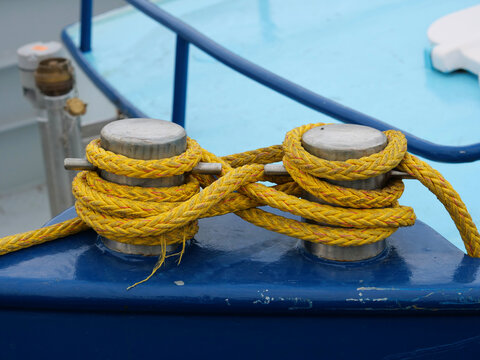 A close-up view of a double stainless steel mooring bollard on a blue painted ship surface, securely wrapped with a thick, braided bright yellow mooring rope in a figure-eight pattern.