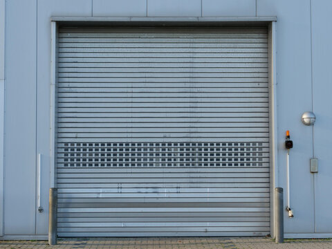 A full front view of a grey metal industrial roller shutter door set into a warehouse facade, featuring a central horizontal ventilation grid section, side bollards, and an outdoor warning light setup