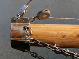 wooden bowsprit of a sailing vessel, showing stainless steel fittings, turnbuckles, a wooden block with rigging rope, and heavy metal chains over the water © Christian Bunge