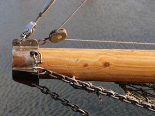wooden bowsprit of a sailing vessel, showing stainless steel fittings, turnbuckles, a wooden block with rigging rope, and heavy metal chains over the water © Christian Bunge