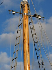a traditional wooden ship mast with visible wood knots, featuring rope ratlines forming a ladder structure and rigging hardware against a blue sky with clouds. © Christian Bunge