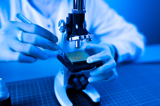 Engineer inspecting semiconductor CPU microchip under optical microscope with tweezers in blue light laboratory